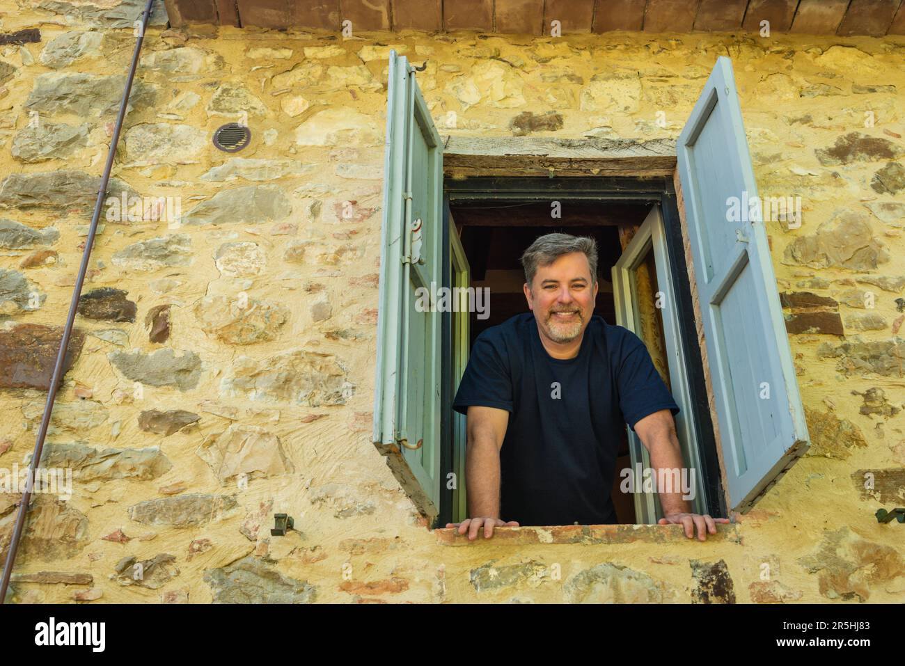 View from outside of man leaning out window from second floor in old stone home with shutters Stock Photo