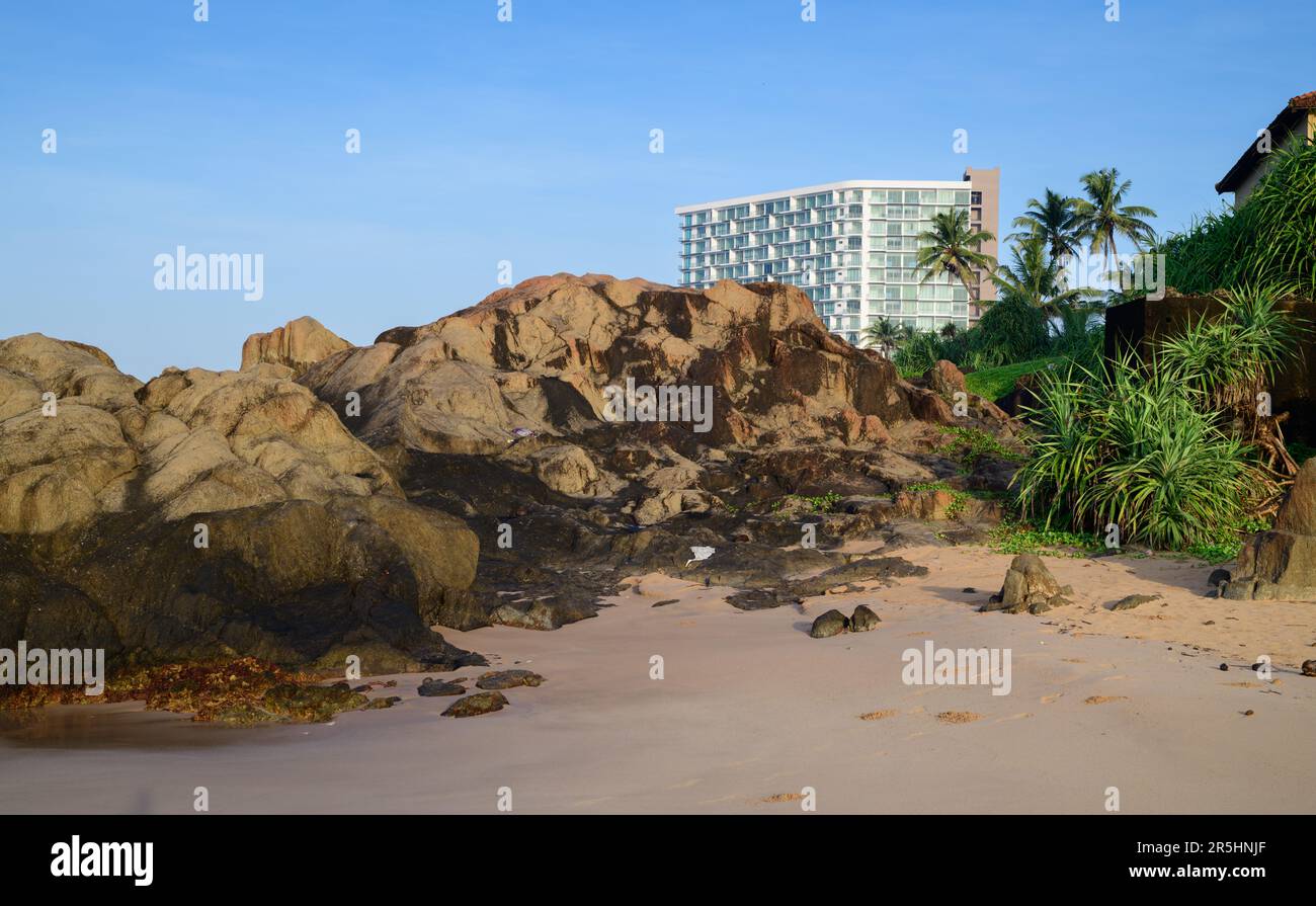 Tropical beach picturesque landscape photograph, rocky sandy beach, and the urban hotel building in the background. Clear blue skies. Stock Photo
