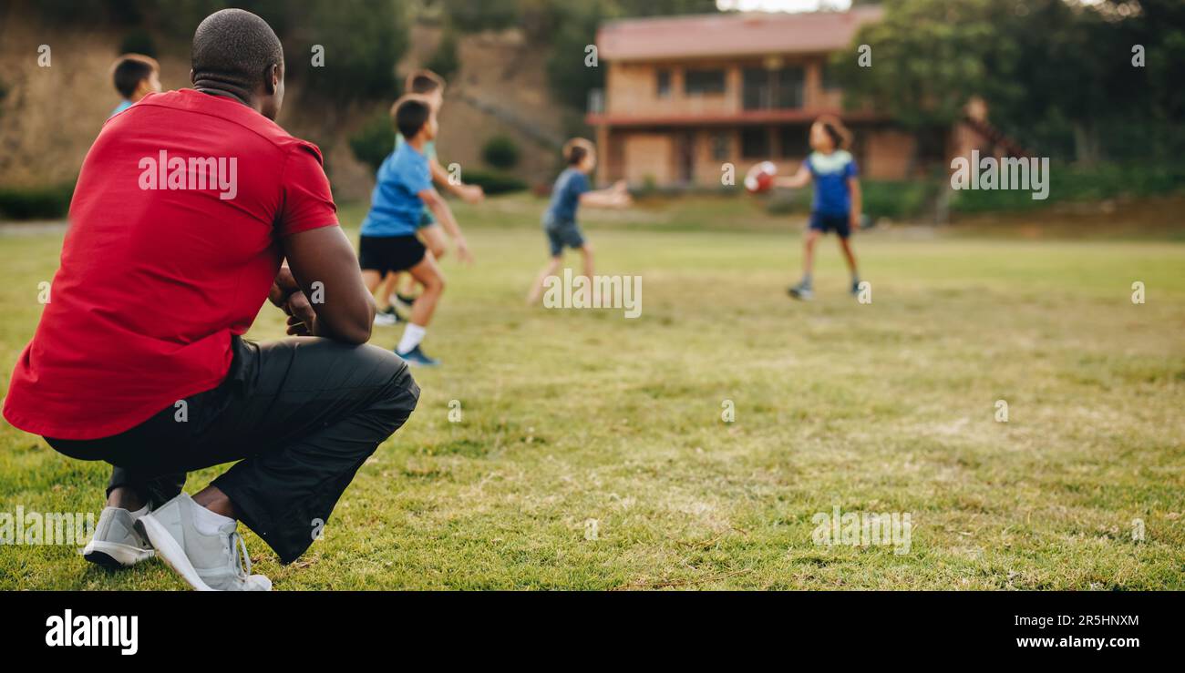 Coach having a practice session with kids in a school. Sports instructor training a group of school children in a rugby field. Sports mentorship in el Stock Photo