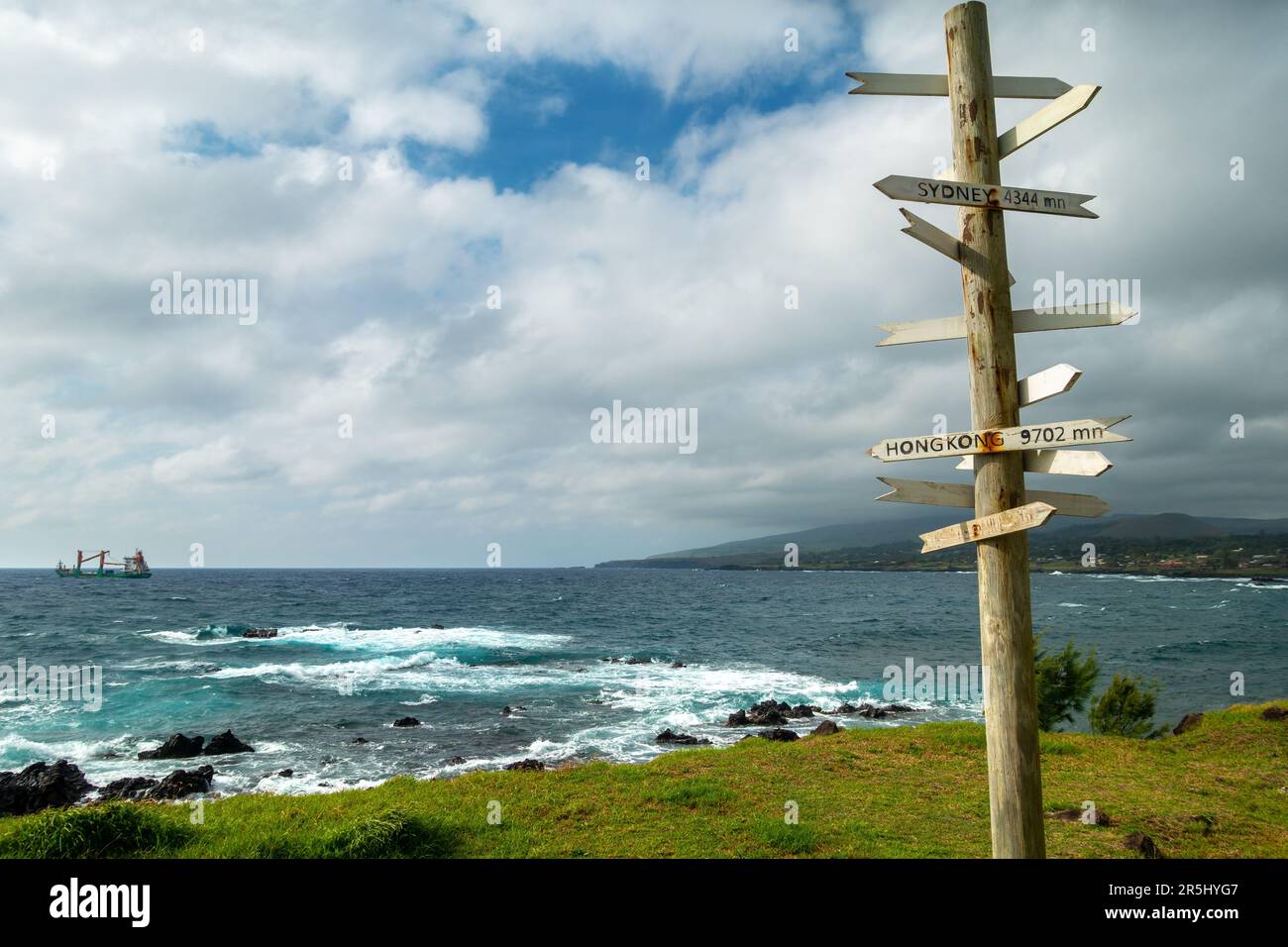 Wooden Post with Direction Signs at Hanga Roa Waterfront.  Easter Island Chile Pacific Ocean Coastline, Stormy Skyline, Distant Cargo Ship on Horizon Stock Photo