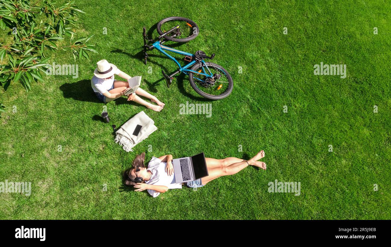 Young girls with bicycle in park, using laptop computer and headphones, two student girls studying online outdoors sitting on grass near bike Stock Photo