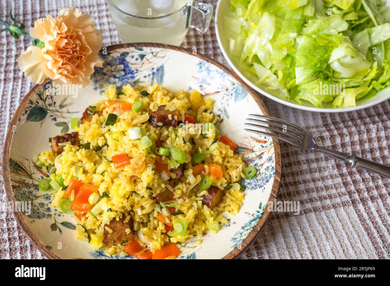 Vegetarian meal. Turmeric rice with green onion, orange pepper, zucchini and tempeh on decorative plate, lettuce salad, flower and drink on table. Stock Photo