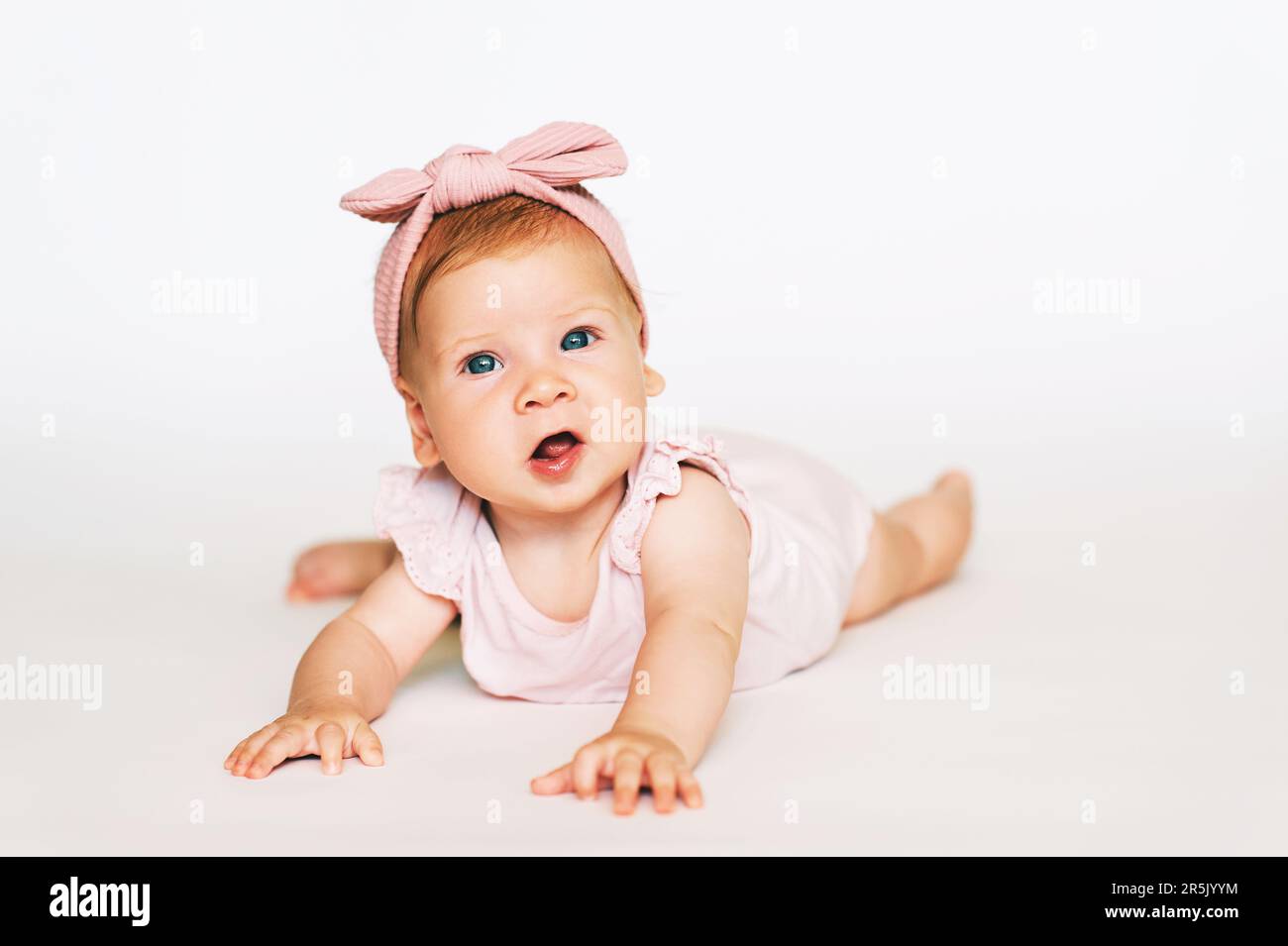 Portrait of adorable red-haired baby lying on belly, white background, wearing pink body and headband Stock Photo