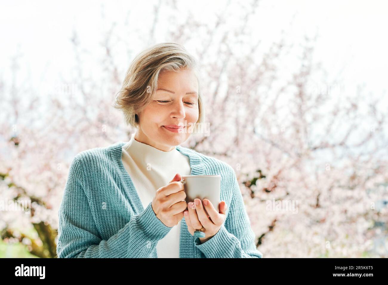 Spring portrait of beautiful and elegant middle age woman posing against pink blooming tree, holding cup of coffee or tea Stock Photo