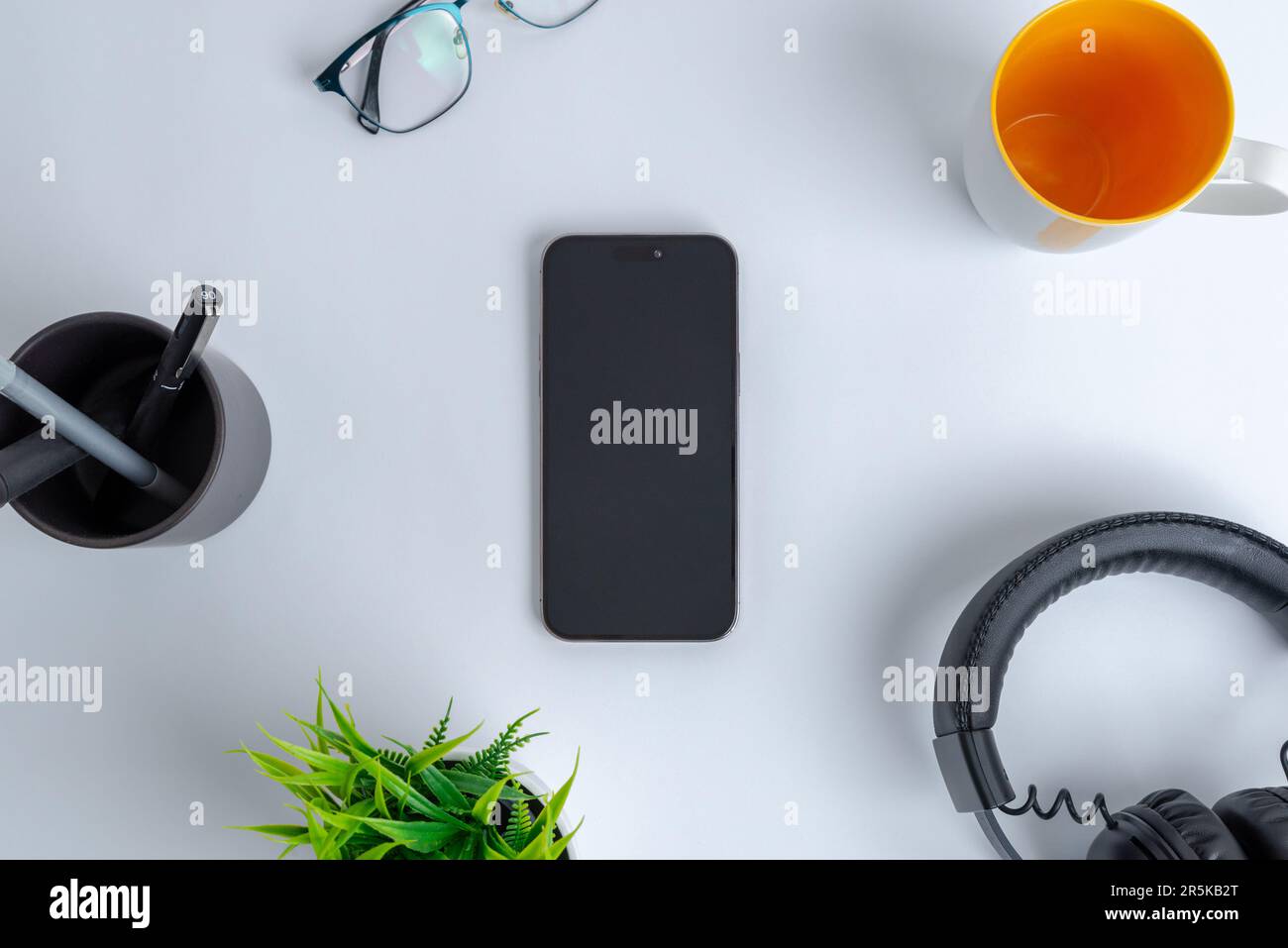 Phone on work desk. Top view, flat lay composition. Surrounded by coffee mug, headset, plant, glasses and pens Stock Photo