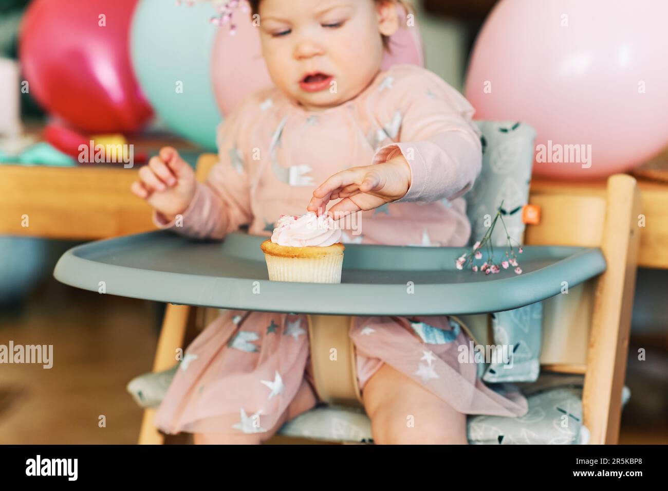 Adorable 1 year old baby girl eating cupcake, happy child sitting in a chair, tasting sweet dessert, first birthday Stock Photo