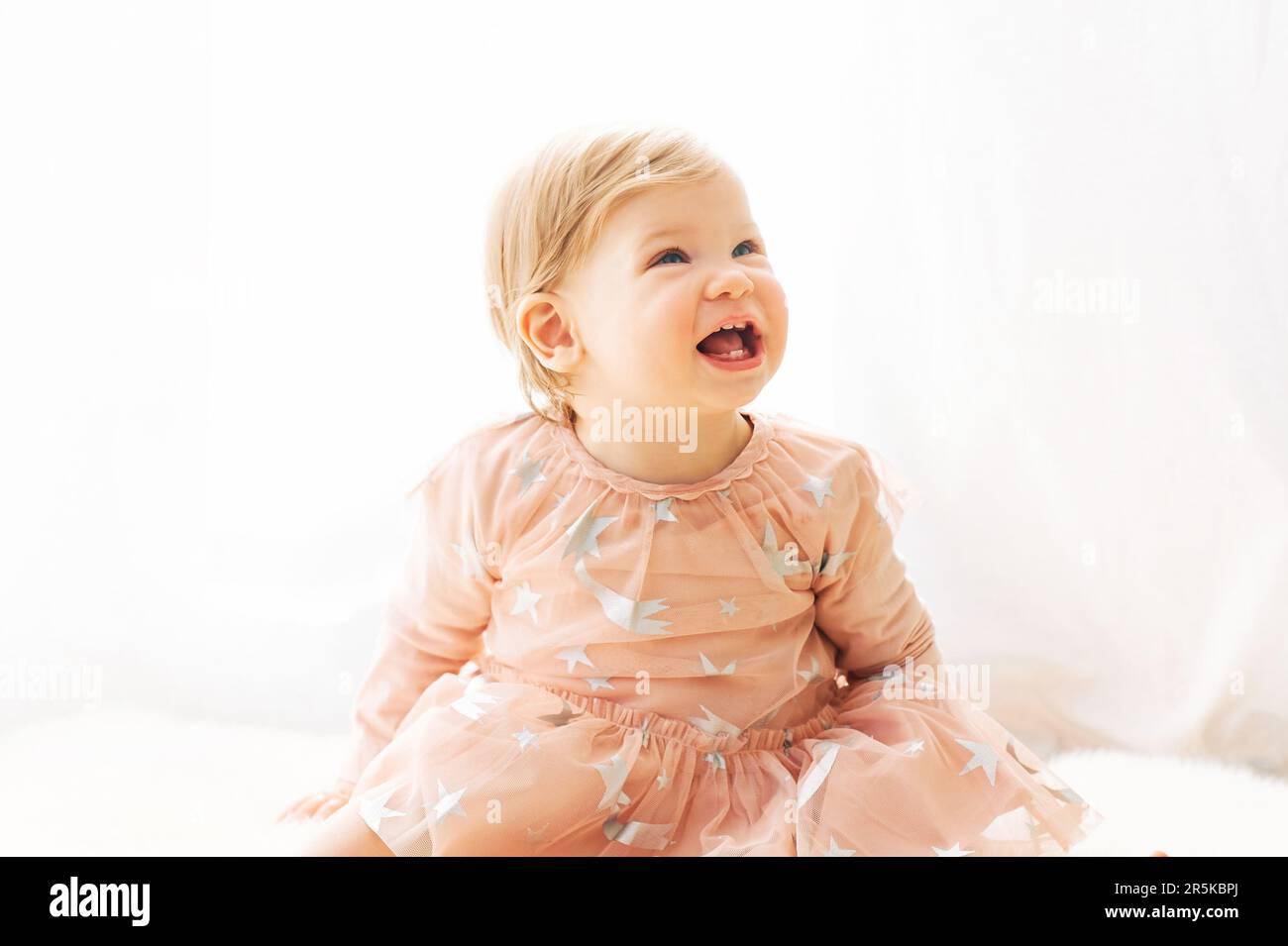 Light soft portrait of cute toddler 1 year old baby girl, wearing pink dress, sitting next to window Stock Photo