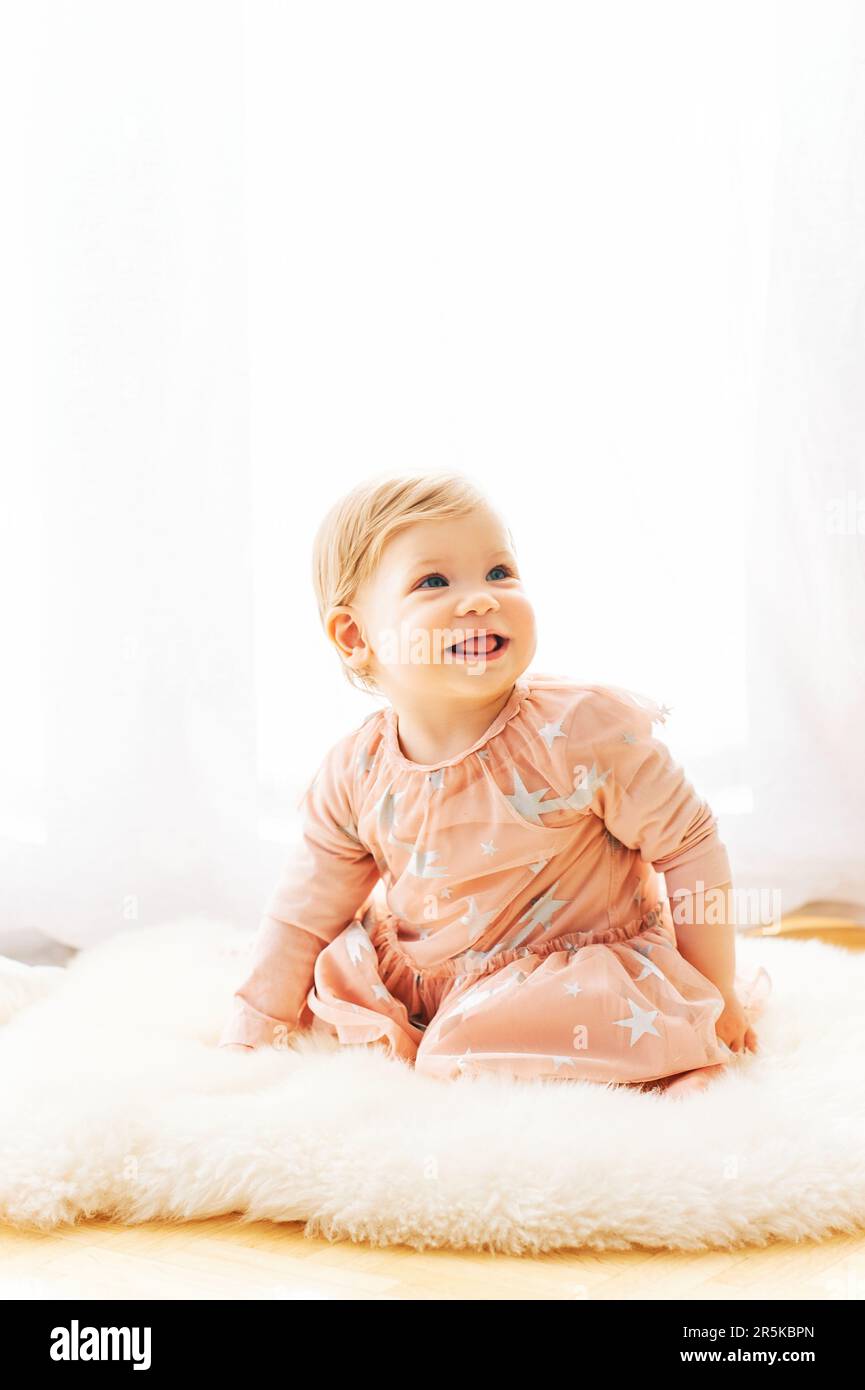 Light soft portrait of cute toddler 1 year old baby girl, wearing pink dress, sitting next to window Stock Photo