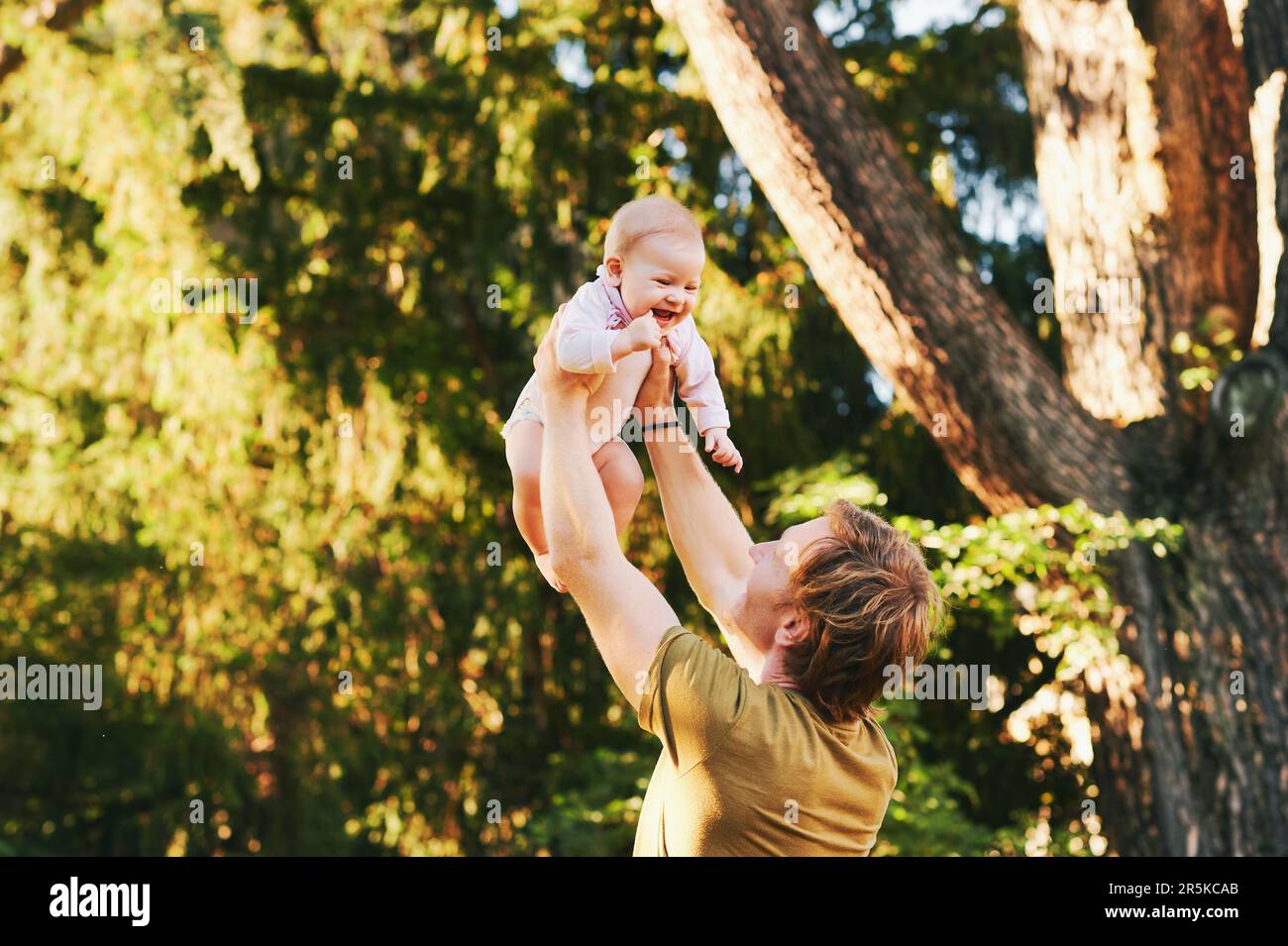 Happy young father playing with adorable baby girl in summer park, family lifestyle, dad holding kid high in the air Stock Photo