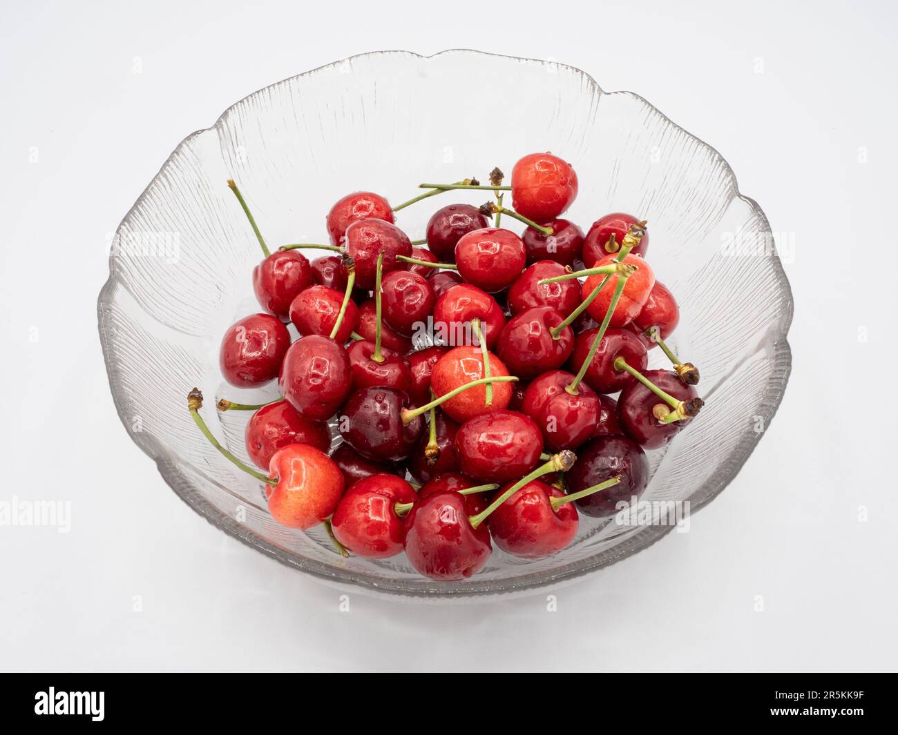 Organic red cherries, glistening with refreshing water droplets, elegantly presented in a pristine glass bowl, perfectly isolated on a clean white bac Stock Photo