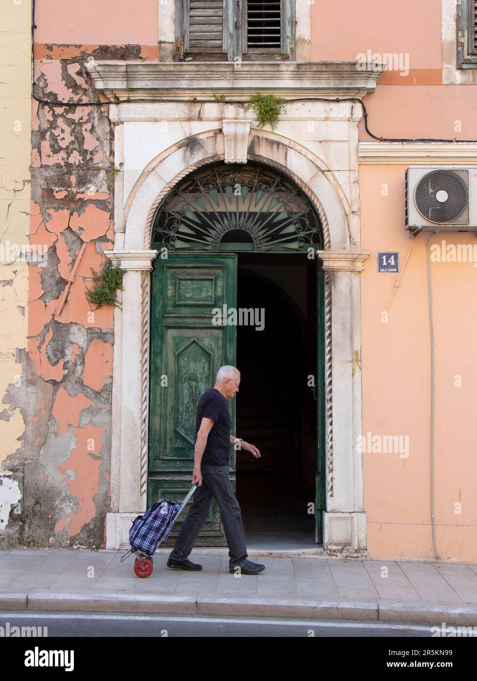 Sibenik, Croatia - May 31, 2023: One elderly man walking by arch entrance door while pulling shopping trolley Stock Photo