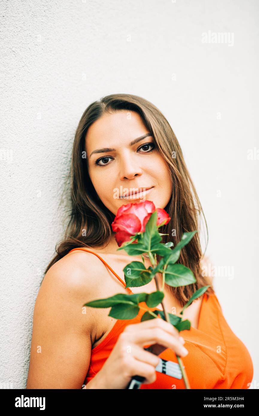 Outdoor portrait of attractive woman holding pink flower, wearing orange clothes Stock Photo
