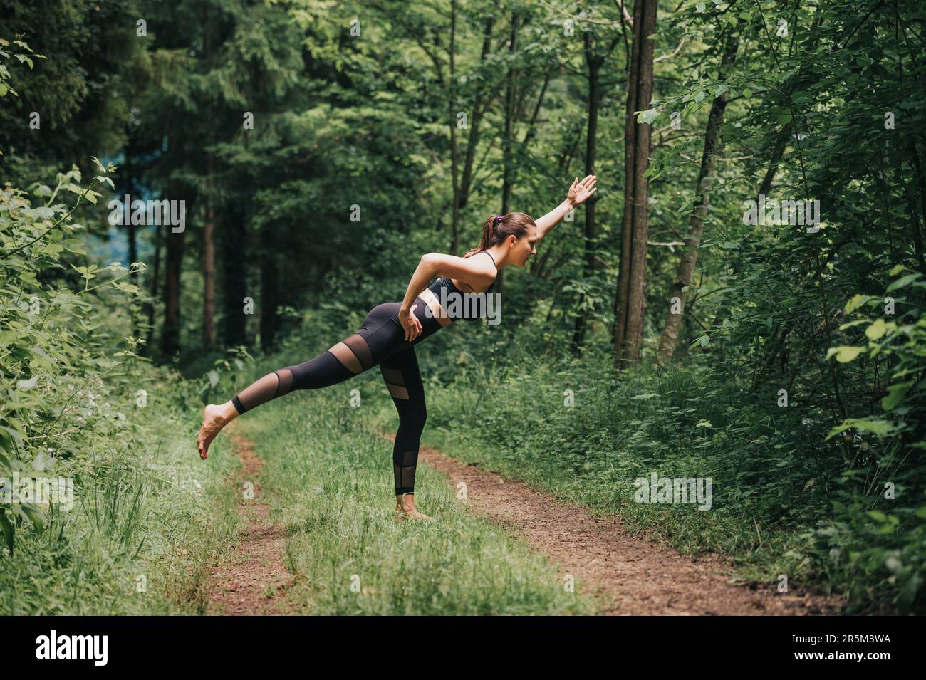 Young fit woman practicing yoga in summer forest, wearing black leggings and top Stock Photo