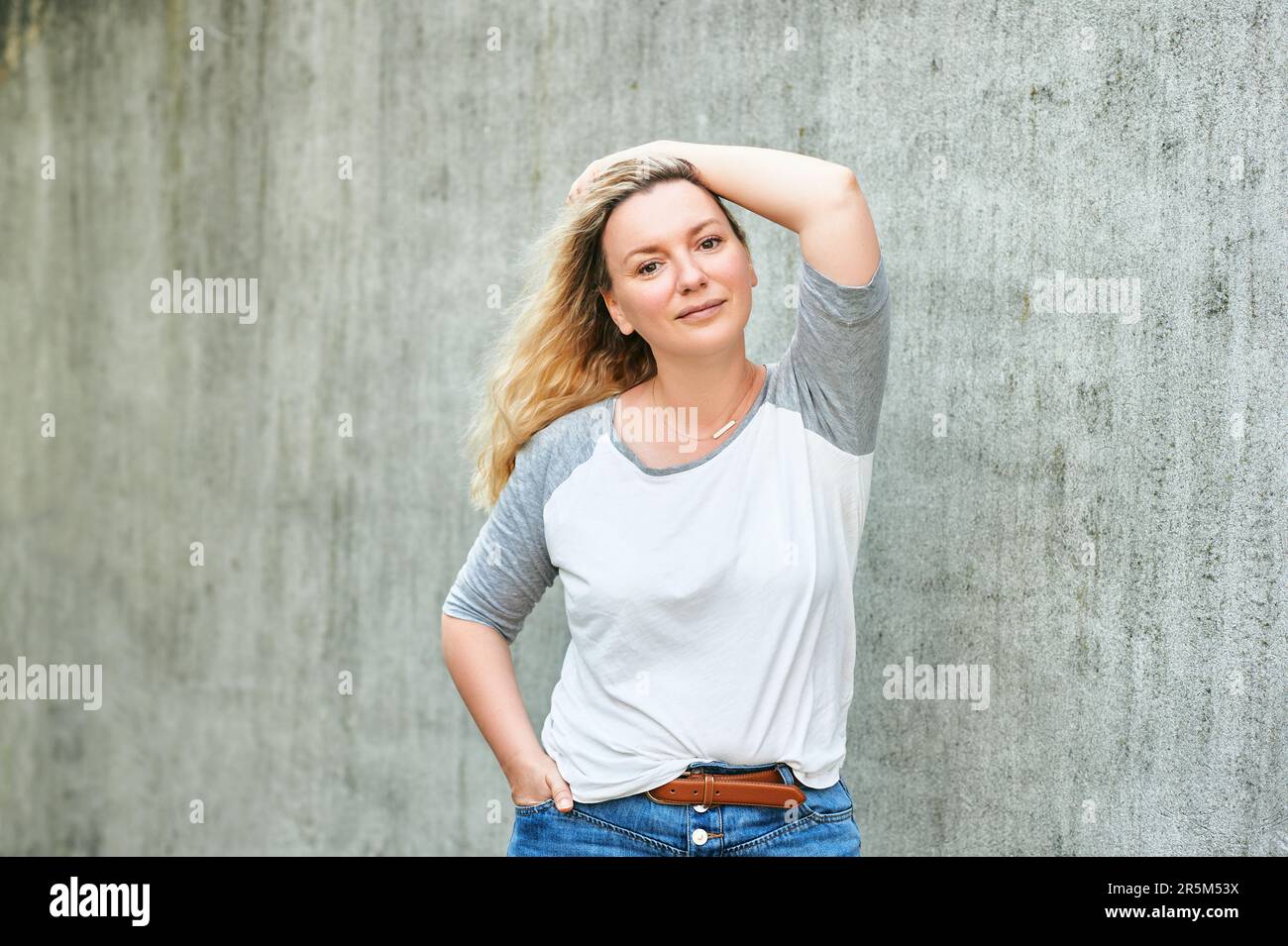 Outdoor portrait of pretty woman posing on grey urban background, wearing white t-shirt with grey sleeves Stock Photo