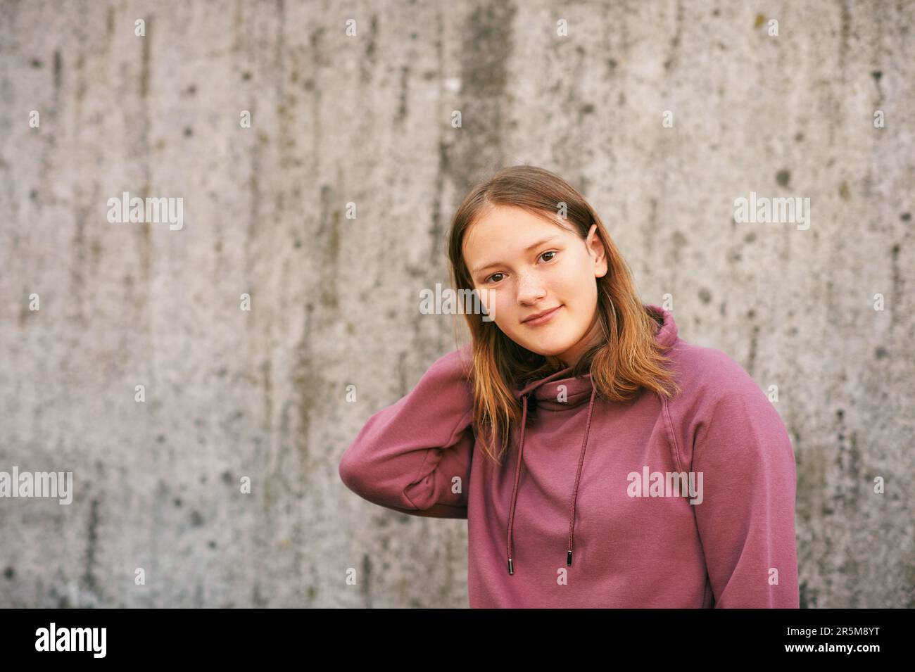 Outdoor urban portrait of young teen girl posing against grey wall, wearing pink sweatshirt Stock Photo