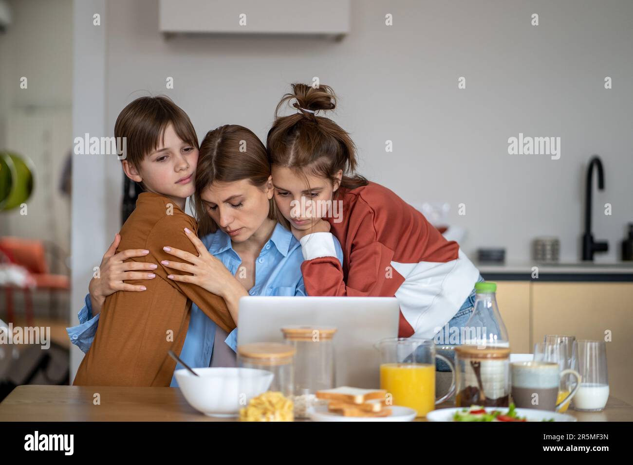 Daughter and son hug mom in kitchen. Sad single mother has problems with online work Stock Photo