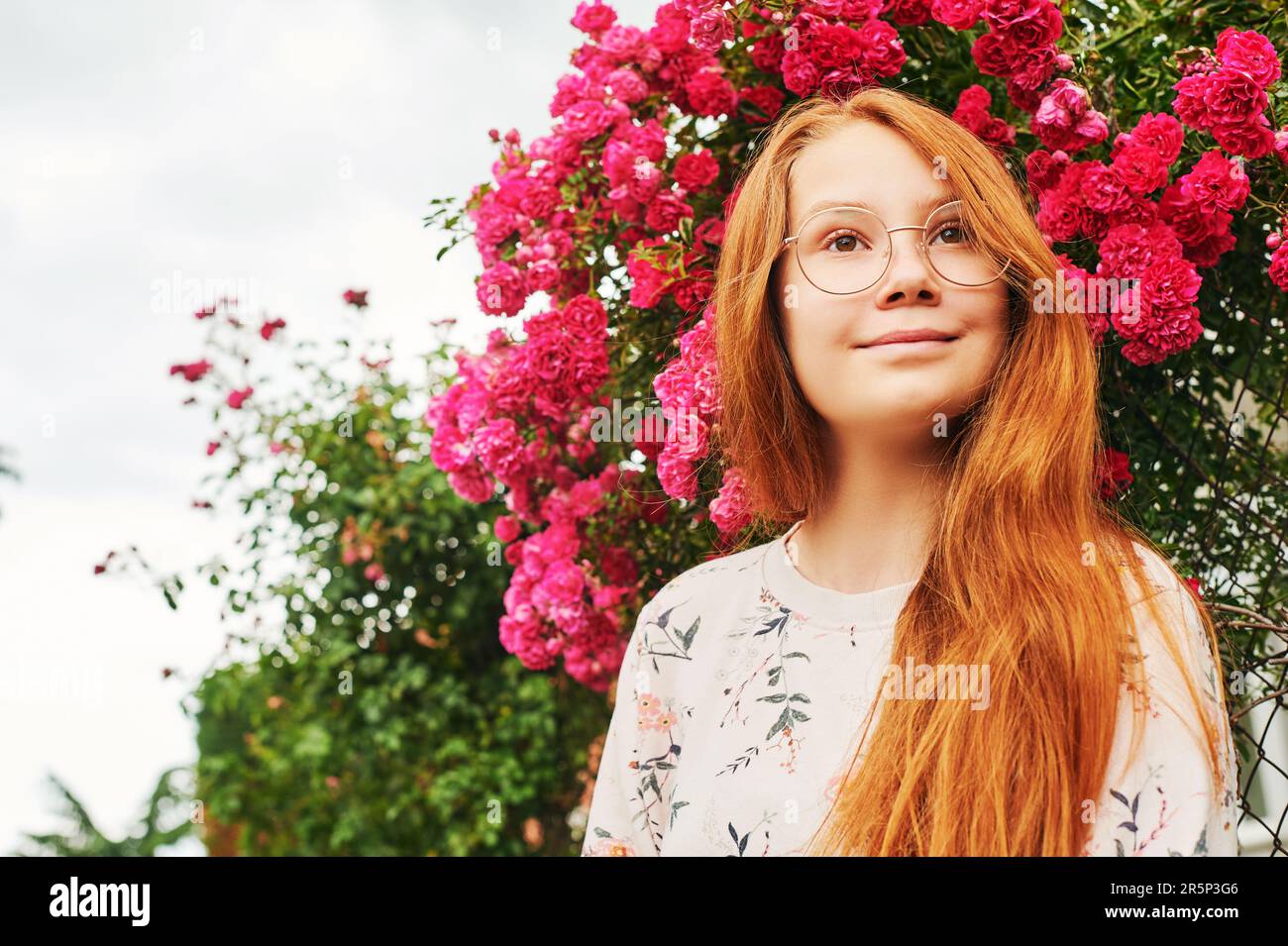 Outdoor close up portrait of happy young girl, wearing eyeglasses, posing in flower garden Stock Photo
