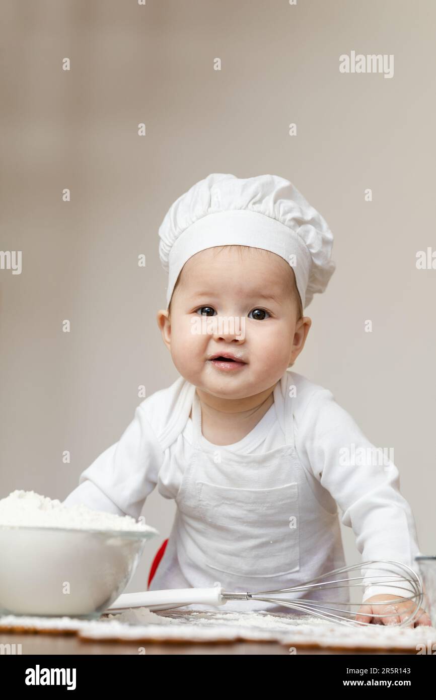 An Asian baby in a chef s cap and apron smeared in flour