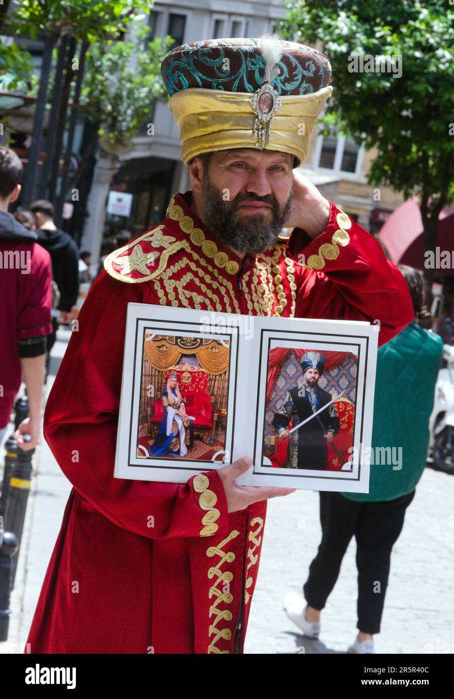 A man in traditional Turkish clothing or clothes or costume stands on the street to try to sell a service in Istanbul, Turkey, the Republic of Türkiye. It appears he is asking tourists to pay to dress in tradional Turkish costume and he will take their photograph to produce a photo print or digital image for them. Stock Photo