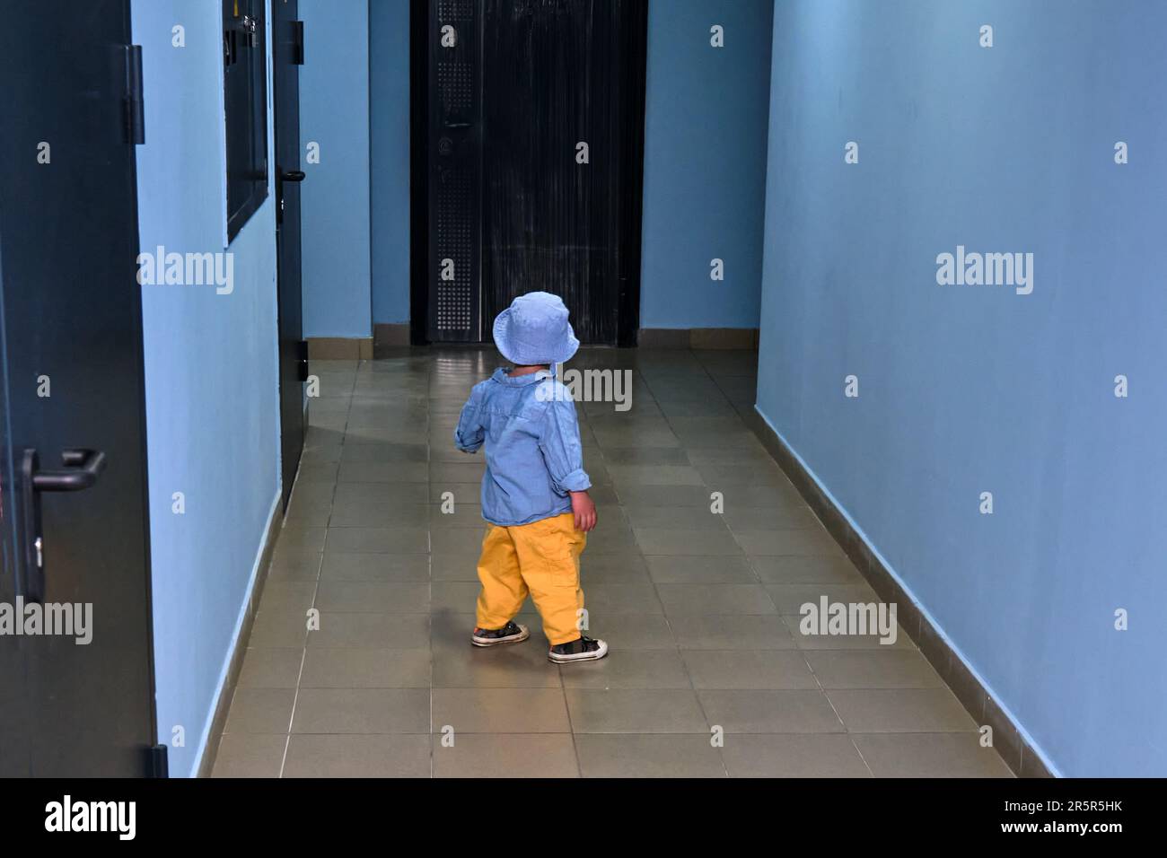 The toddler baby walks in the entrance hall on the floor near the apartment doors. A child runs in the hallway of an apartment building. Kid aged abou Stock Photo