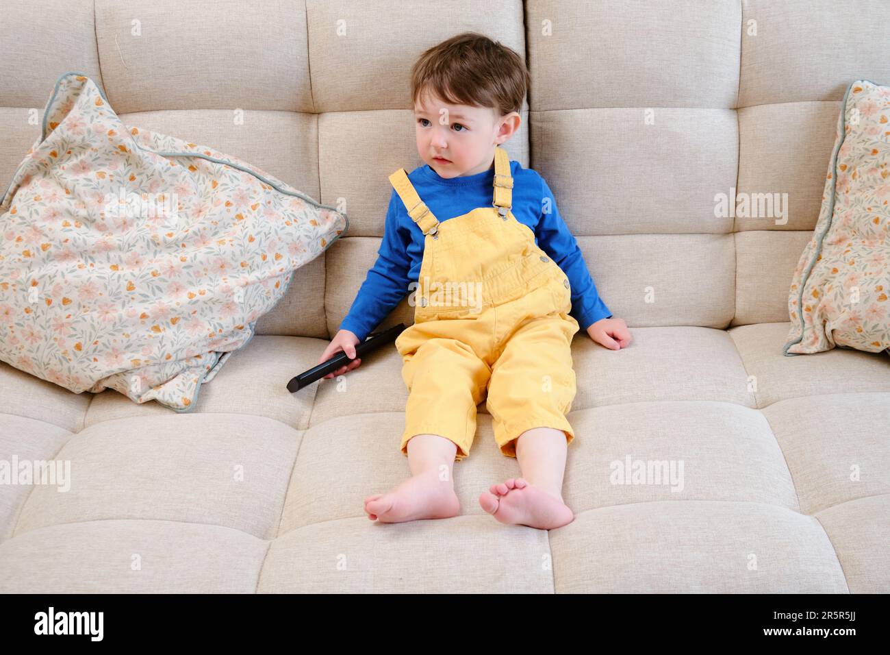 The little child sits on the sofa, watching TV with a remote control in hand and a smile on their face. The happy baby is watching their favorite show Stock Photo