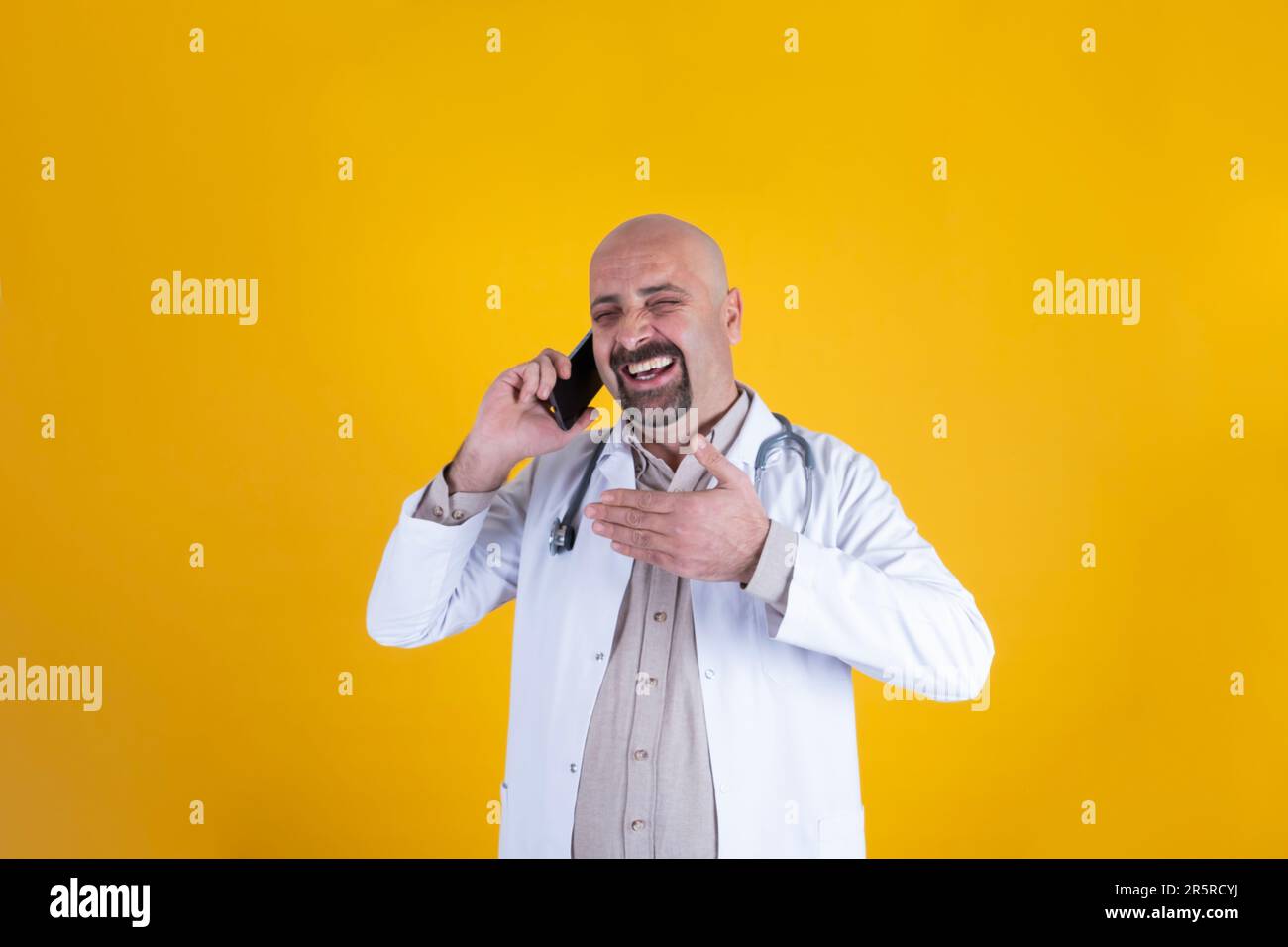 Bald caucasian male doctor talking on the phone. Happy physician wearing hospital uniform, stethoscope. Lol. Speaking with friend, cheerful face. Stock Photo