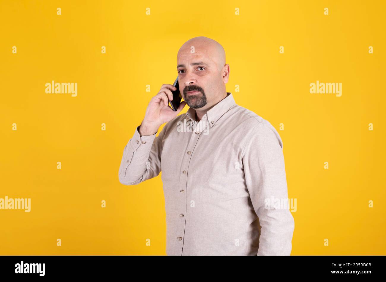 Standing over yellow studio background. Bald male seriously listening call. Businessman talking with client. Copy space. Stock Photo