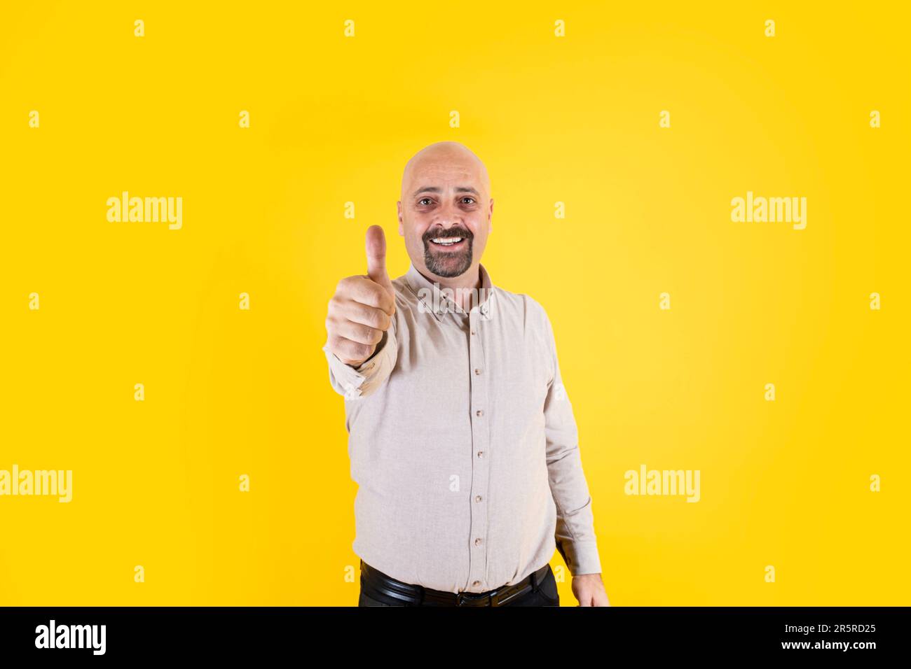 Middle aged bald man doing happy thumbs up gesture approving expression. Wearing casual beige shirt isolated over yellow background, copy space. Stock Photo