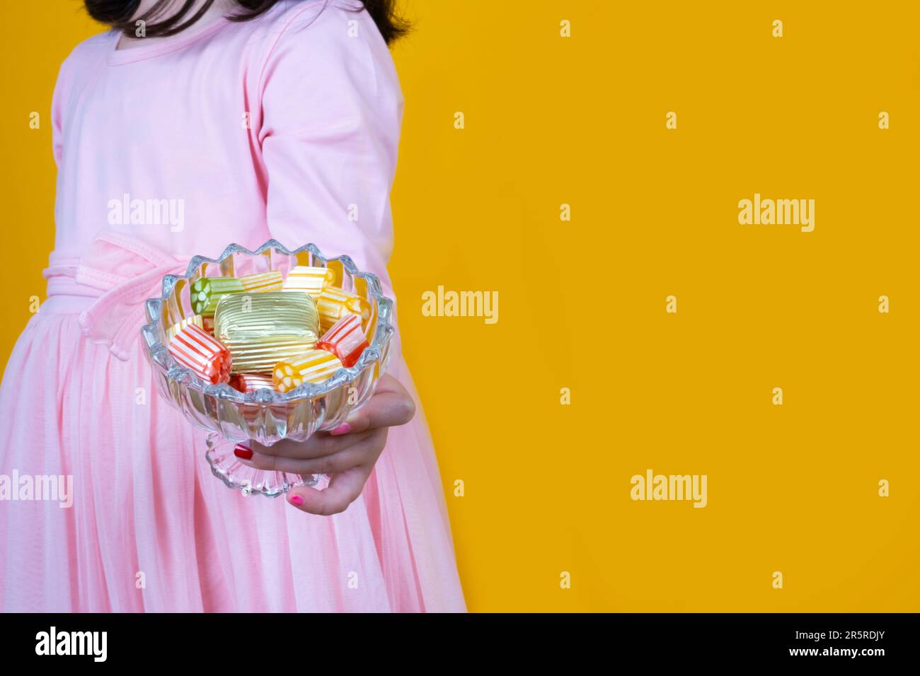 Little girl holding bowl of wrapped luxury chocolate. Standing over isolated yellow background, copy space. Traditional religious holiday sugar feast Stock Photo