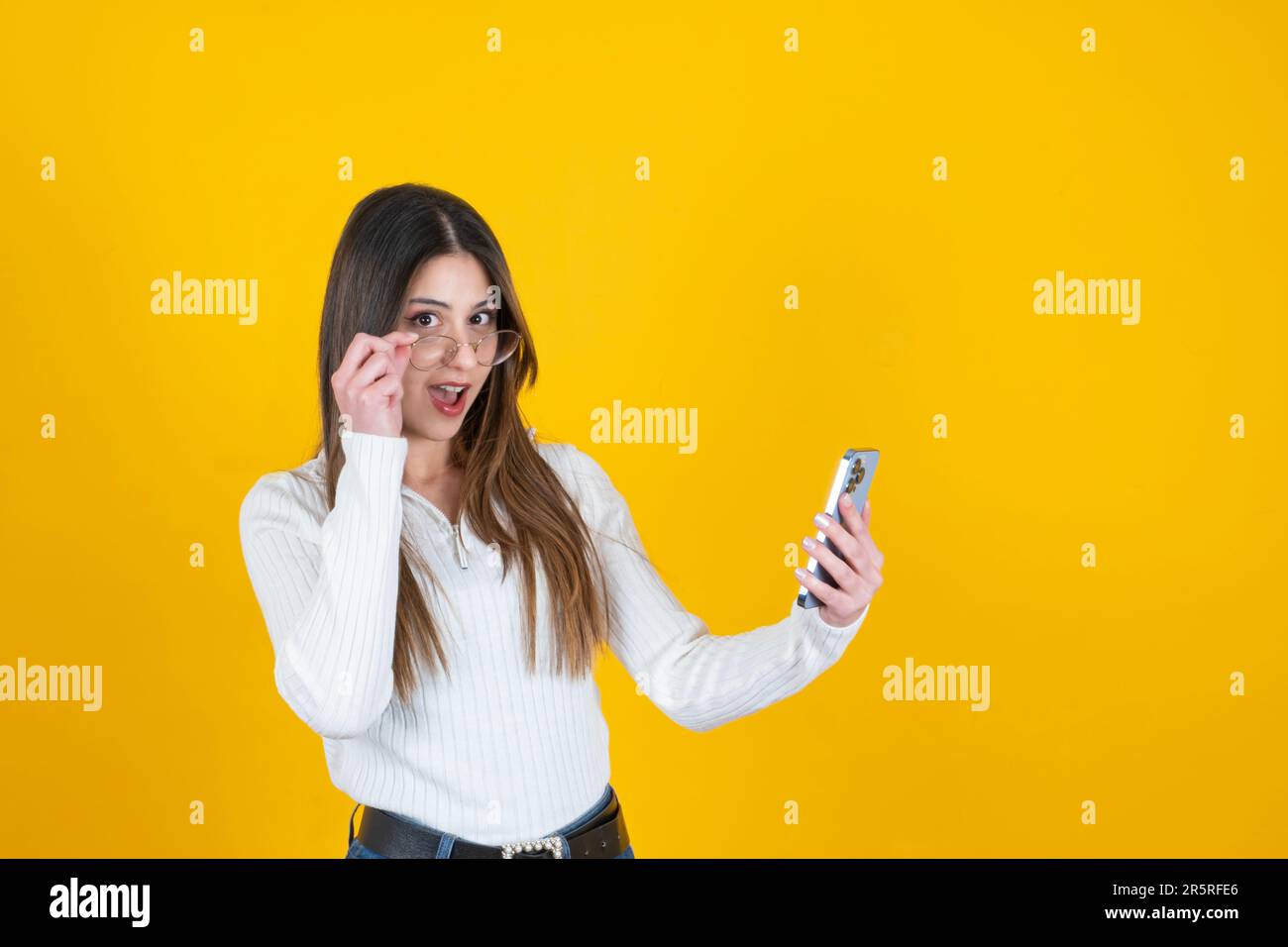 Surprised young woman. Shocked 20s caucasian girl looking smartphone and under glasses with open mouth. Isolated yellow orange background. Copy space Stock Photo