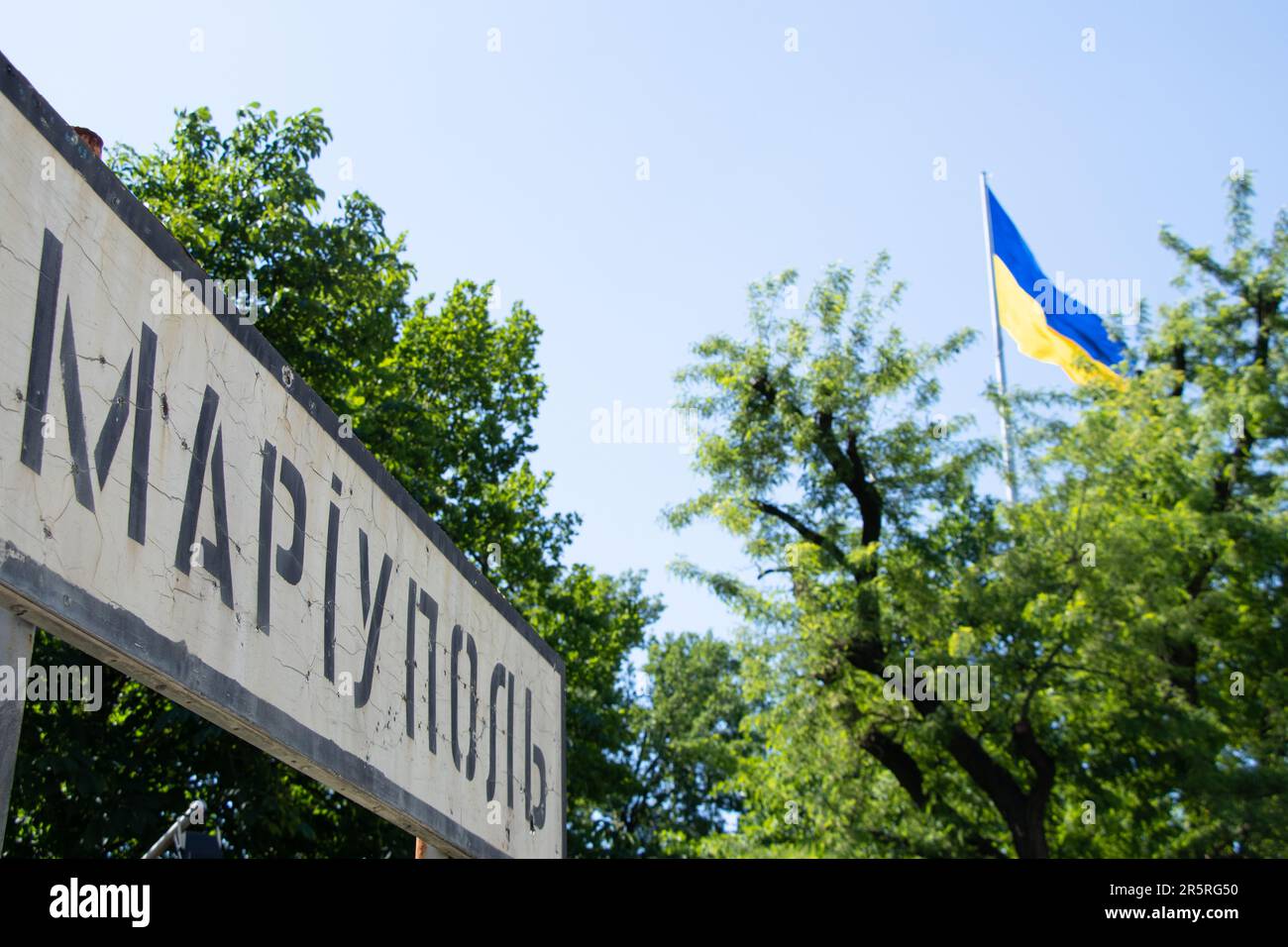 The name of the city in Ukrainian Mariupol on a road sign broken from bullets against the background of the flag of Ukraine in the sky, an occupied ci Stock Photo