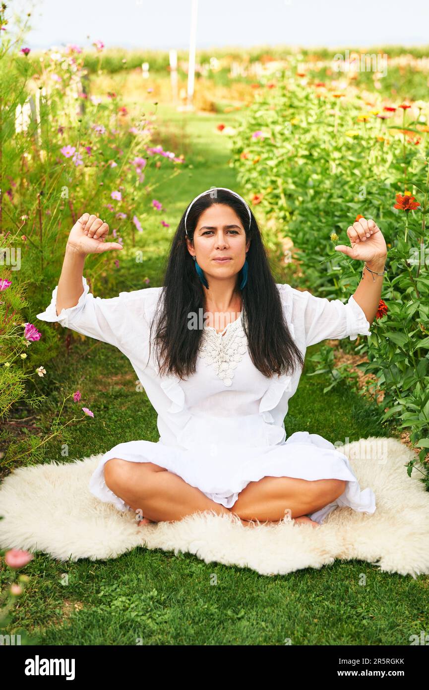 Middle age woman meditating in green garden, sitting on sheep skin Stock Photo