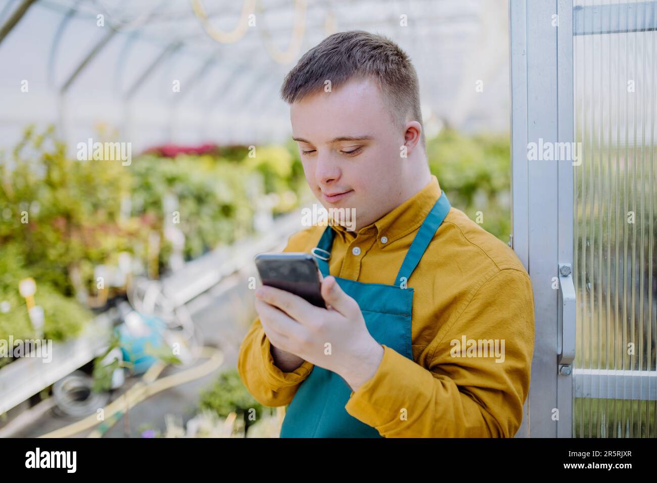 Young employee with Down syndrome working in garden centre, standing in door of greenhouse with cellphone. Stock Photo