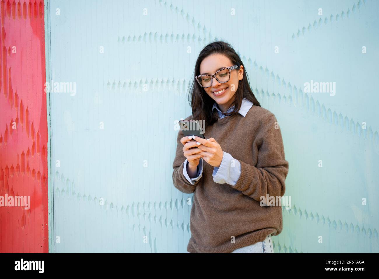 Satisfied hipster girl wear glasses, types text message on mobile phone,  enjoys online communication, types feedback, wears sweater, isolated on  blue color background. Technology concept Stock Photo - Alamy