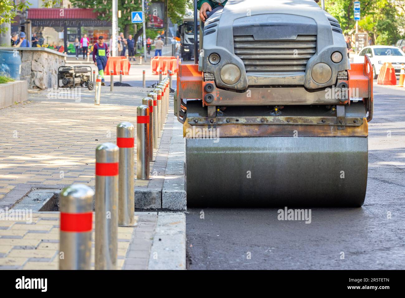 A vibrating roller compacts and compacts fresh asphalt on a city street on a sunny day on a roadway along a paved sidewalk. Close-up. Copy space. Stock Photo