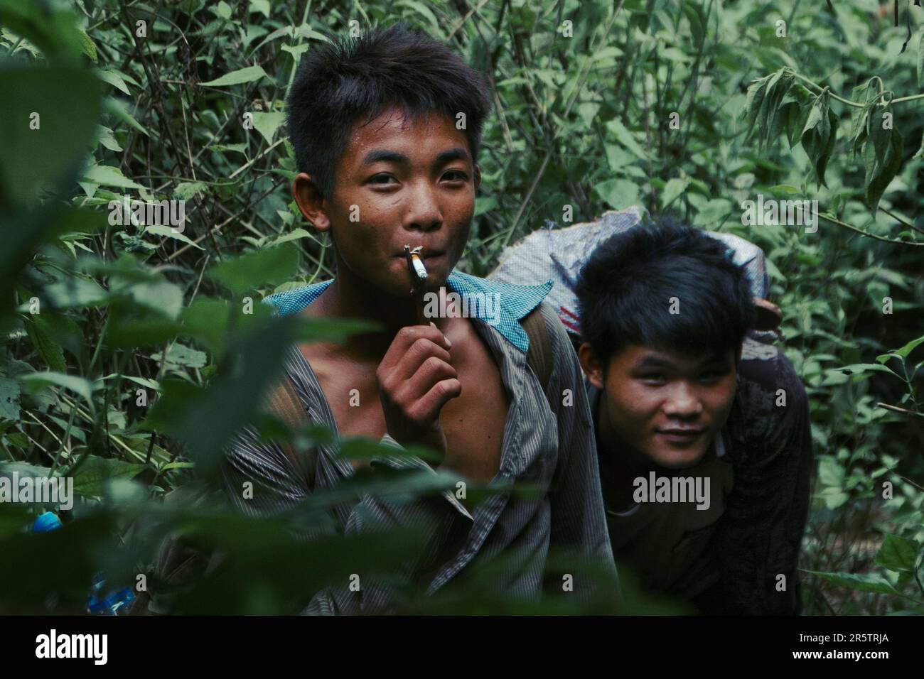 Two young boys, friends, living in the moment, enjoying a cigarette, while being engulged and framed by the wild jungle. Stock Photo