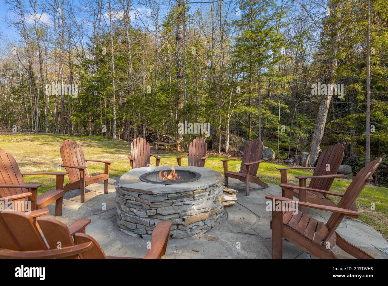 A cozy outdoor setting featuring four wooden chairs placed around a fire pit constructed with natural stone Stock Photo