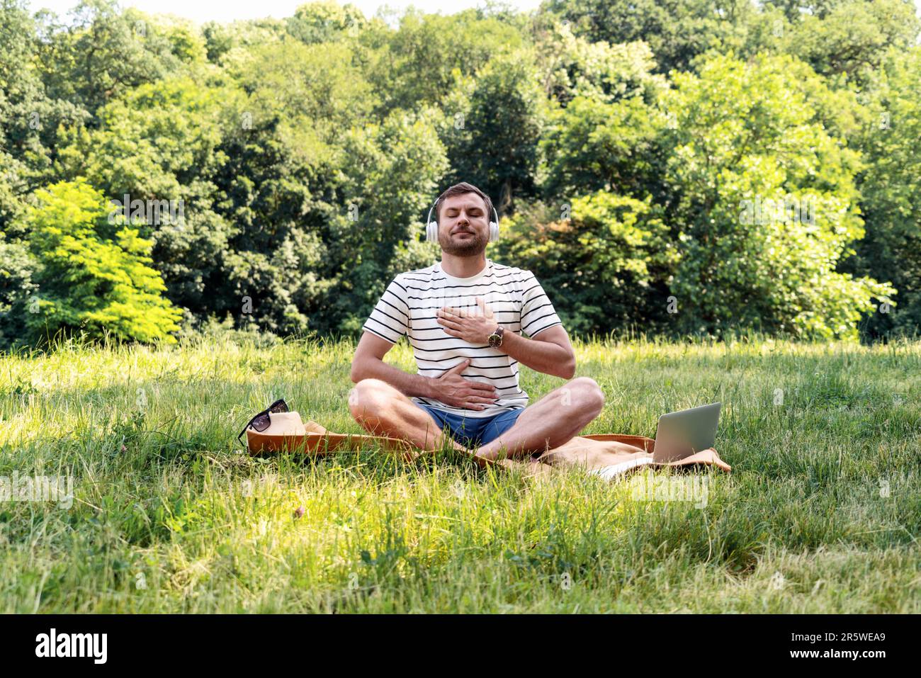 Man doing yoga breathing exercise sitting on the grass in city park in summer, listening to meditation music. Stock Photo