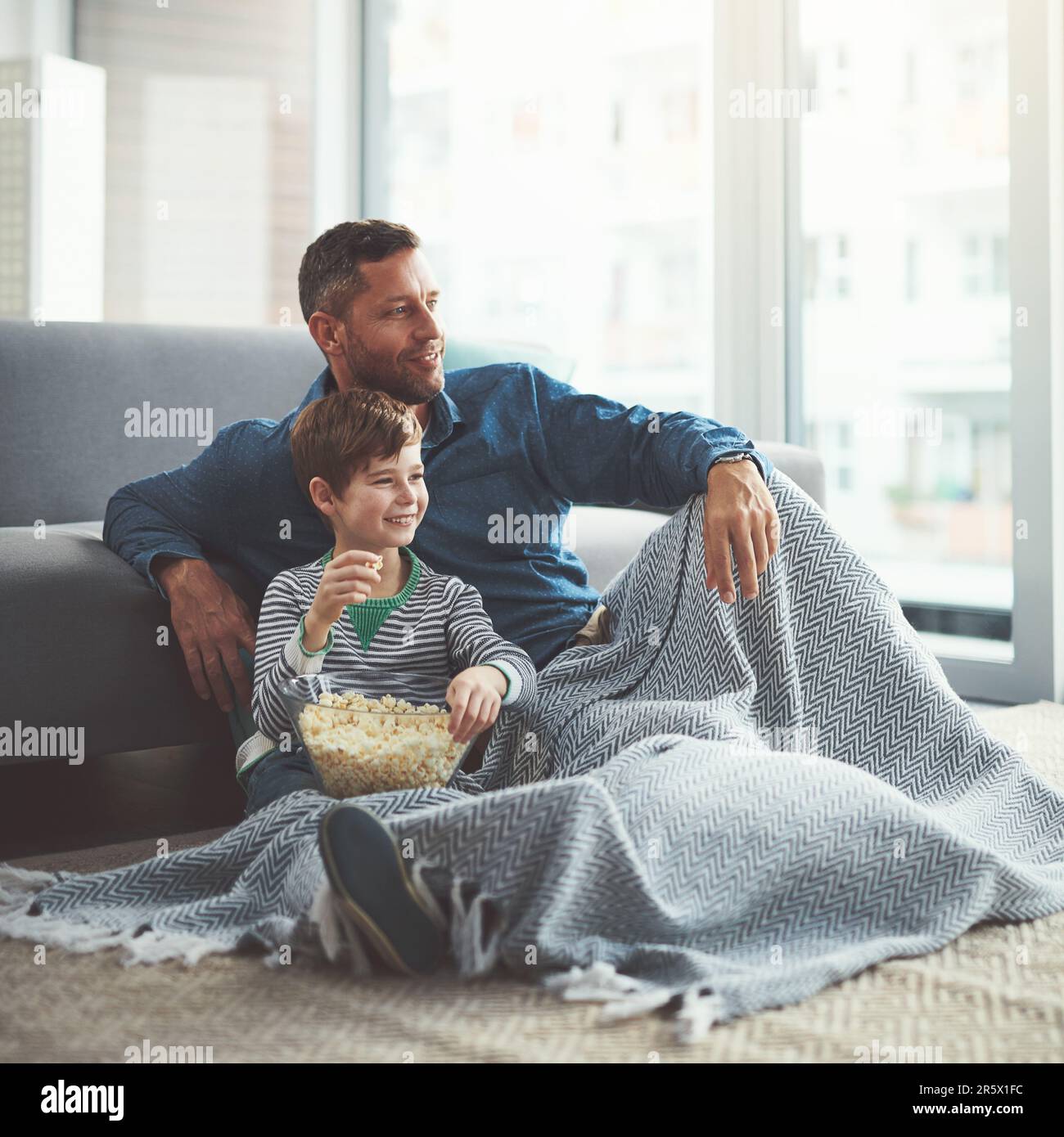 Thanks for the popcorn dad. a carefree young boy and his father watching a movie together while being seated on the floor at home during the day. Stock Photo