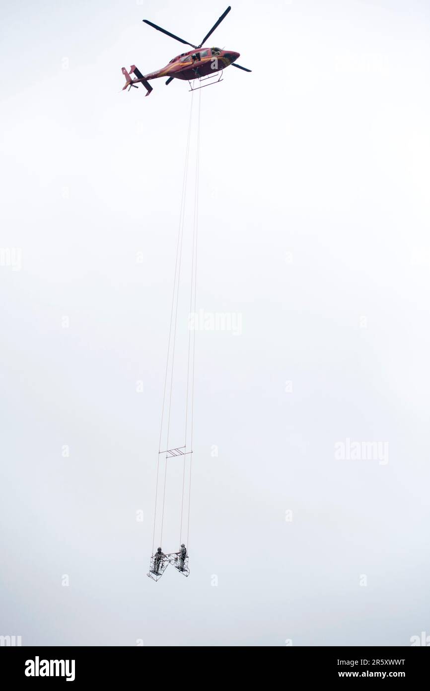 WUHU, CHINA - JUNE 6, 2023 - A helicopter uses a basket to 'lift' two power maintenance personnel onto a defective ultra-high voltage line by the Yang Stock Photo