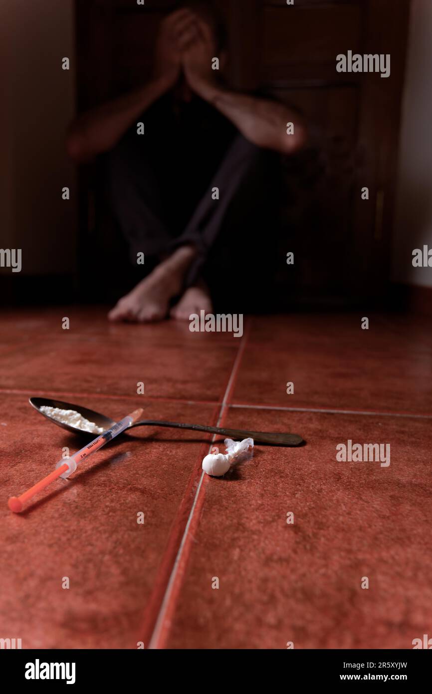 Man lying on the floor in the dark with his hands on his head desperate for drugs in the foreground Syringe with heroin and spoon Stock Photo
