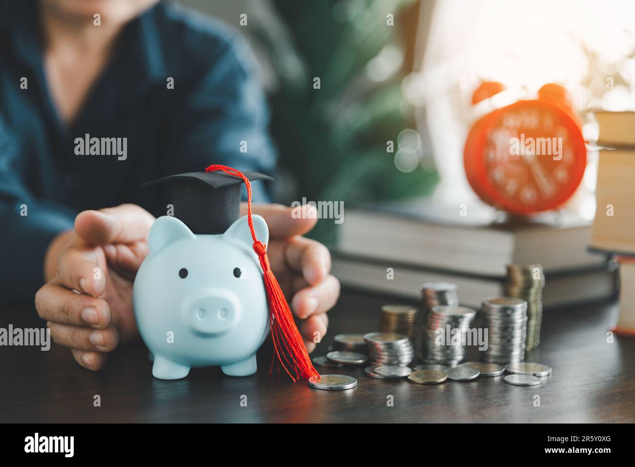 Hands protecting graduation cap on piggy bank with stack coins. Concept of saving money for scholarship to study abroad at university level. Financial Stock Photo