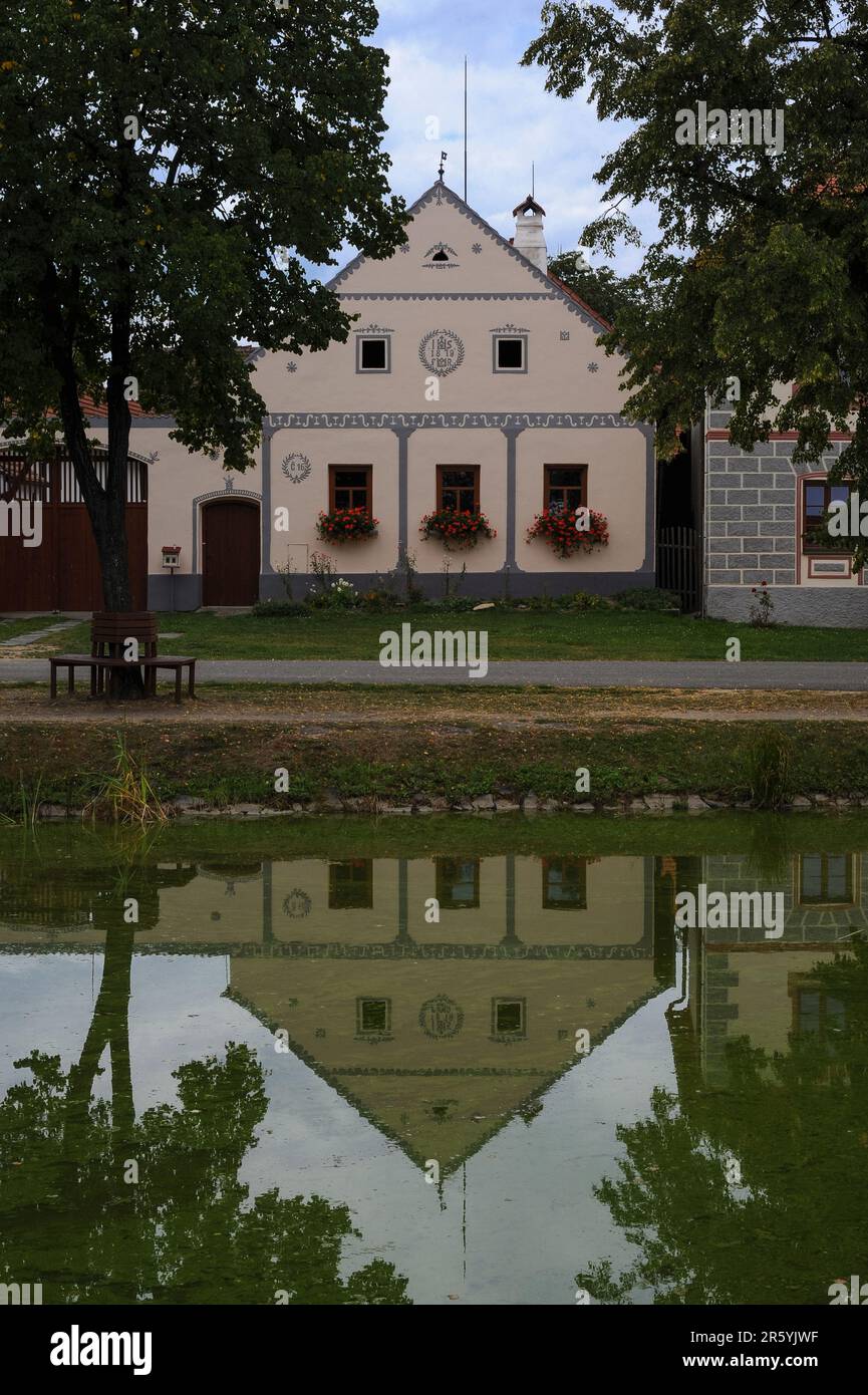 Facade of farmstead in Holašovice, South Bohemia, Czech Republic or Czechia, reflected in the still water of a pond on the village green.  Holašovice is a well-preserved example of a traditional Central European village with a ground plan dating from the Middle Ages and a number of 1700s AD and 1800s AD vernacular buildings in a style known as South Bohemian Folk Baroque, Rural Baroque or Rustic Baroque.  In 1997 the village was included in the UNESCO World Cultural Heritage List. Stock Photo