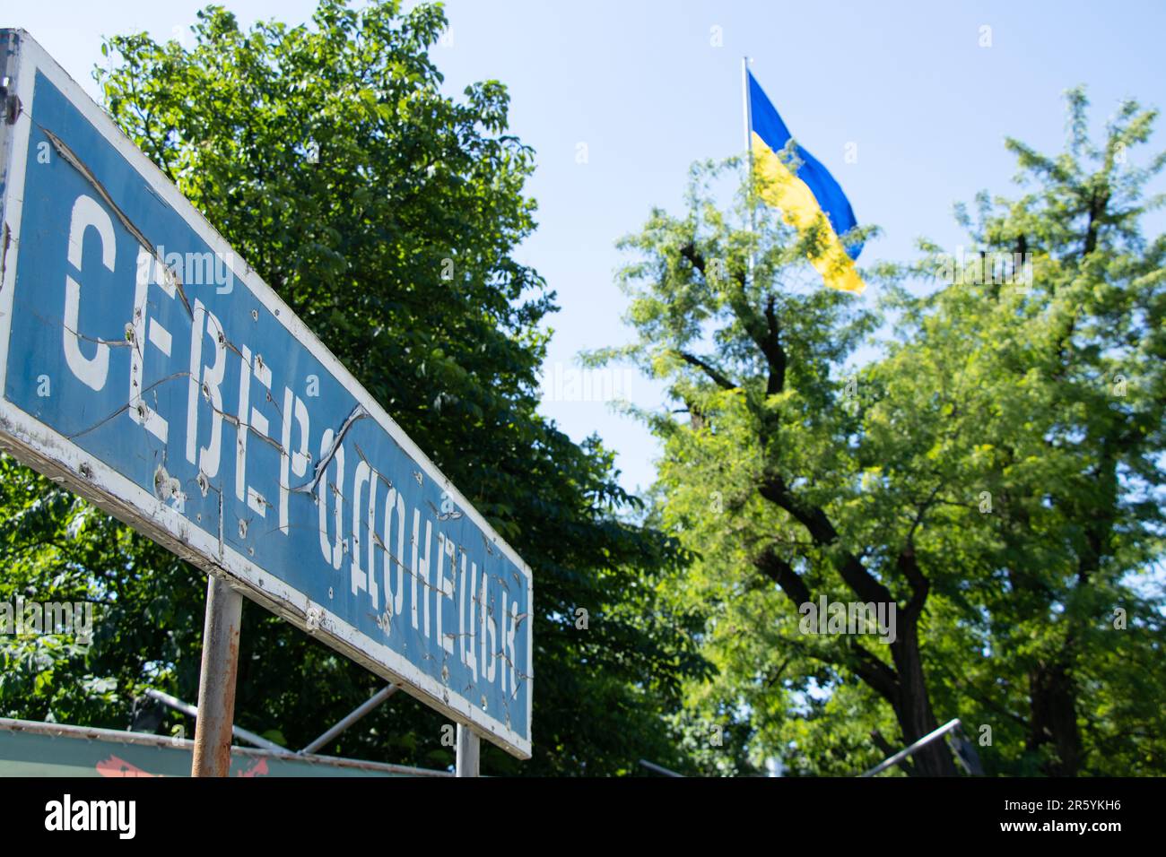 The name of the city in Ukrainian Severodonetsk on a road sign broken from bullets against the background of the flag of Ukraine in the sky, a city in Stock Photo