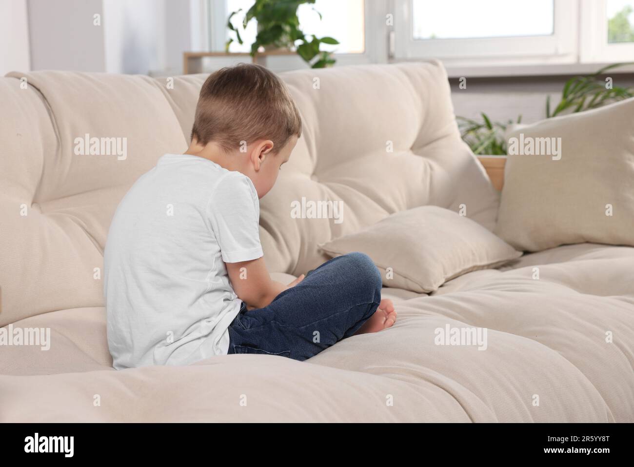 Boy with poor posture using phone on beige sofa in room. Symptom of scoliosis Stock Photo