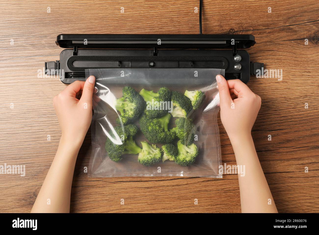 Woman using vacuum sealer at wooden table, top view. Green broccoli in