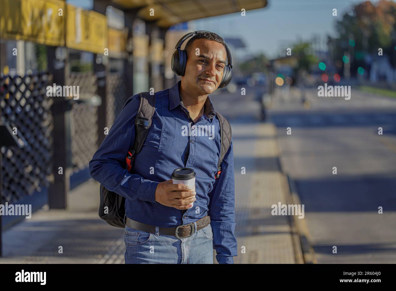 Young latin man with headphones waiting for the bus with a paper cup of coffee in hand. Stock Photo
