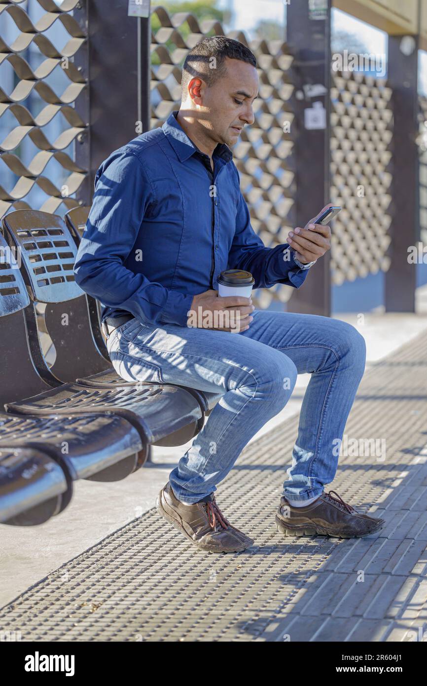 Young latin man sitting waiting for the bus and looking his mobile phone. Stock Photo