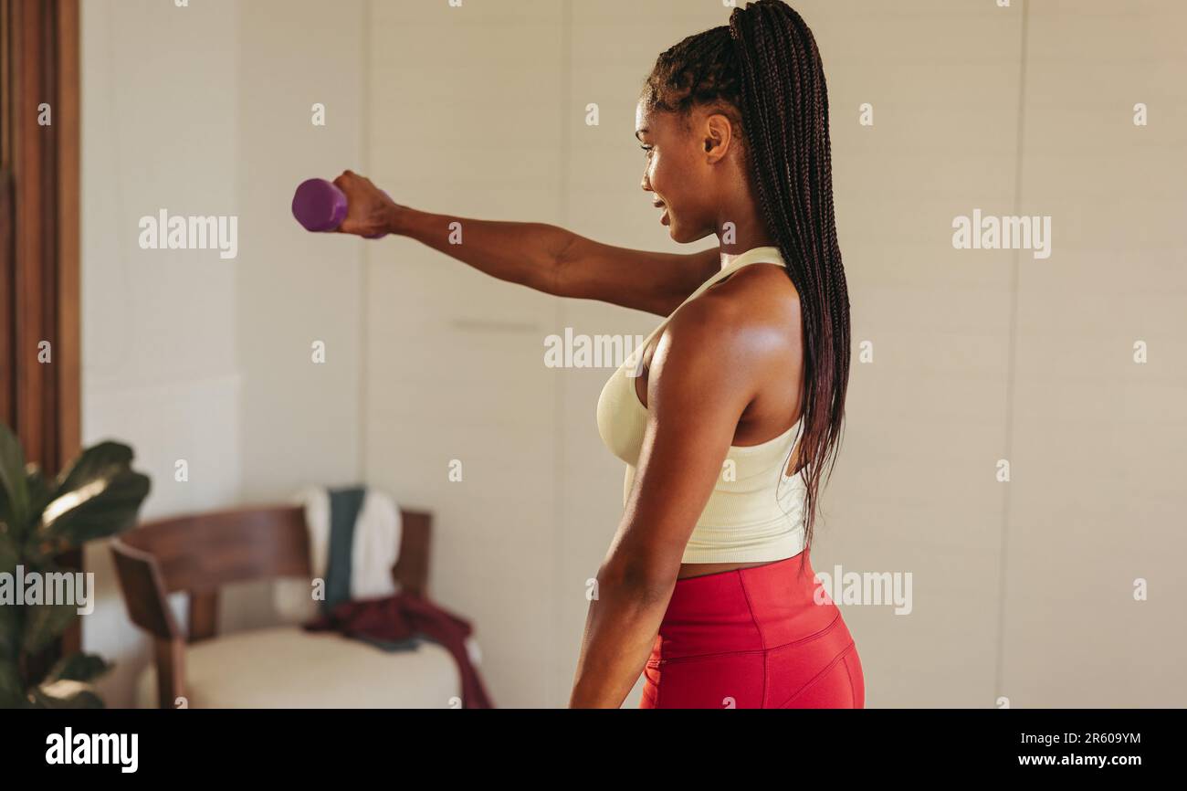 Young black woman working out with dumbbells in her home gym, focusing on strength training as part of her fitness routine. Female athlete crushing he Stock Photo
