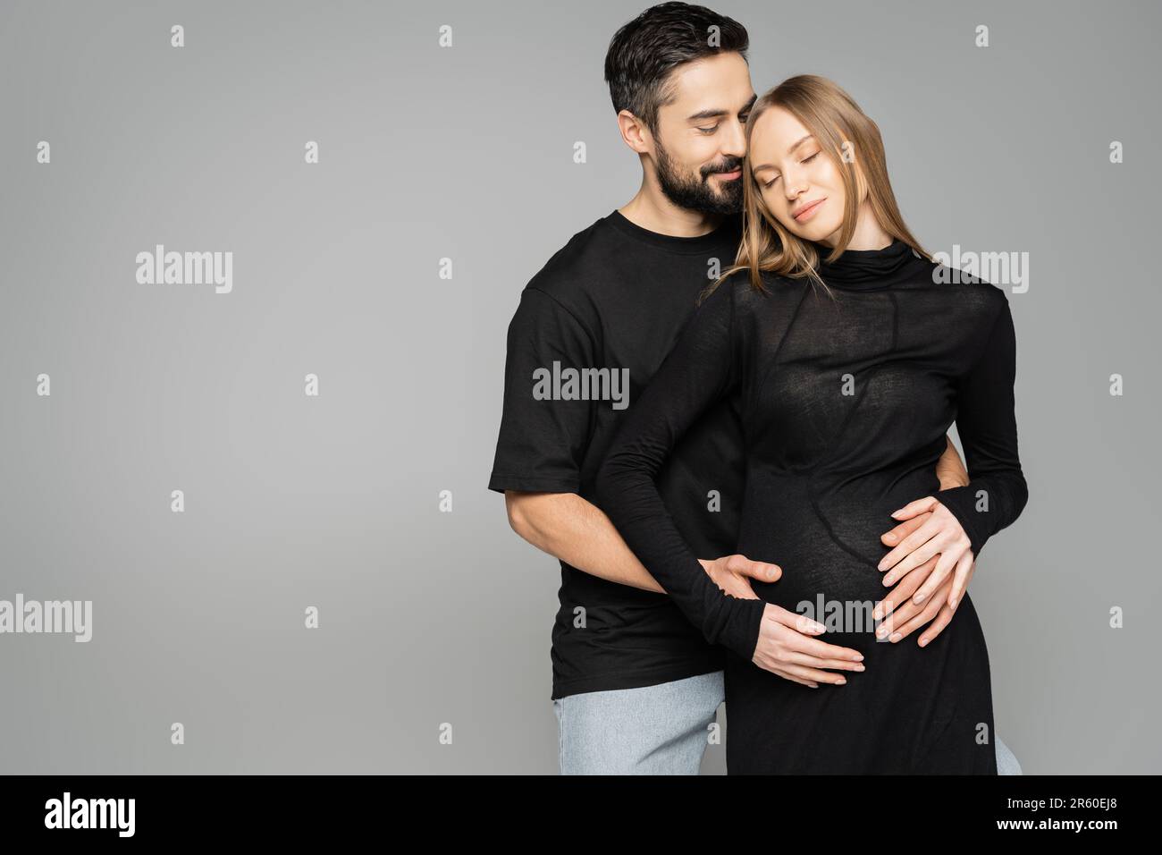 Positive bearded husband in black t-shirt hugging stylish pregnant wife in dress and closing eyes and standing isolated on grey, new beginnings and an Stock Photo